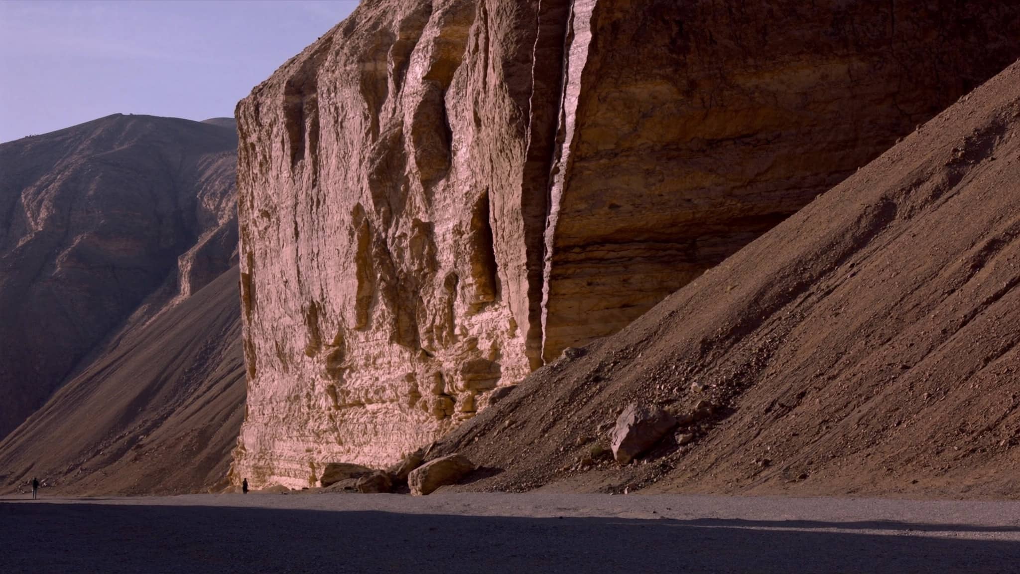 A tall eroded desert escarpment rising above a dry gravel plain, with two figures at its base for scale